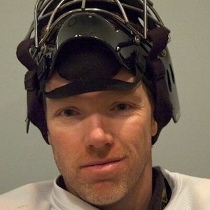 Hockey goalie wearing a wire cage mask and baseball cap liner in a close-up portrait shot