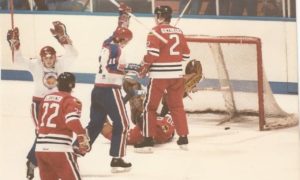 Goalie sprawled on ice after allowing a goal in vintage international hockey game, red and white team players react nearby