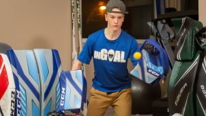 Young goalie in InGoal blue t-shirt holding a tennis ball among CCM leg pads and goalie gear in an equipment room