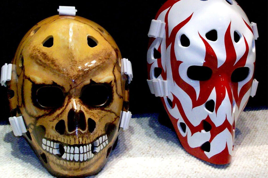 Two Halloween-themed goalie masks: a realistic skull design and a red demon face on white fiberglass cage-style masks