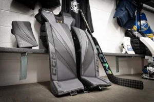 CCM Axis XF goalie leg pads and blocker displayed in a hockey locker room stall with stick and Buffalo Sabres jersey visible