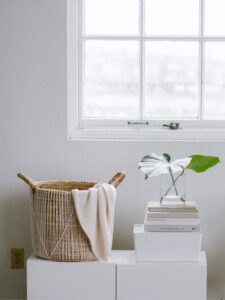 Wicker basket with cream blanket beside stacked books and plant vase on white dresser near bright window
