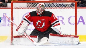 Photo illustration of Connor Hellebuyck in a New Jersey Devils jersey and white TRUE pads, set in his butterfly in front of the net